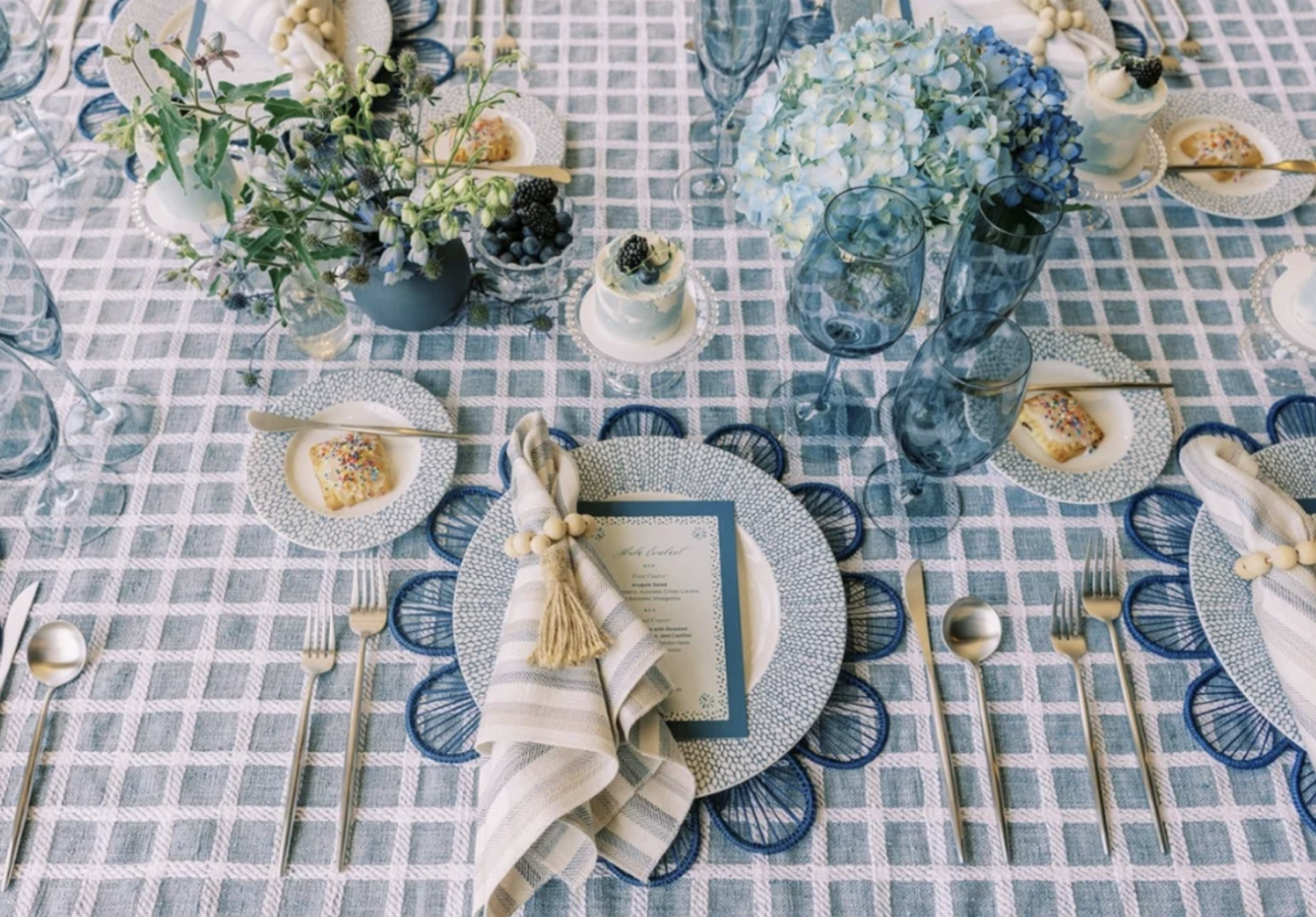 A neatly set table with a blue and white checkered tablecloth, featuring plates, cutlery, glasses, a menu, cloth napkins, and centerpieces of flowers and greenery inspired by summer wedding color trends.